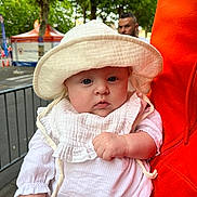 Lylia participe au concours pour gagner de l'argent avec cette photo : adult, baby, blurred_background, child, cute, daylight, expressionless, face, hand, hat, holding, infant, orange_jacket, outdoor, person, portrait, street, trees, urban, white_clothing
