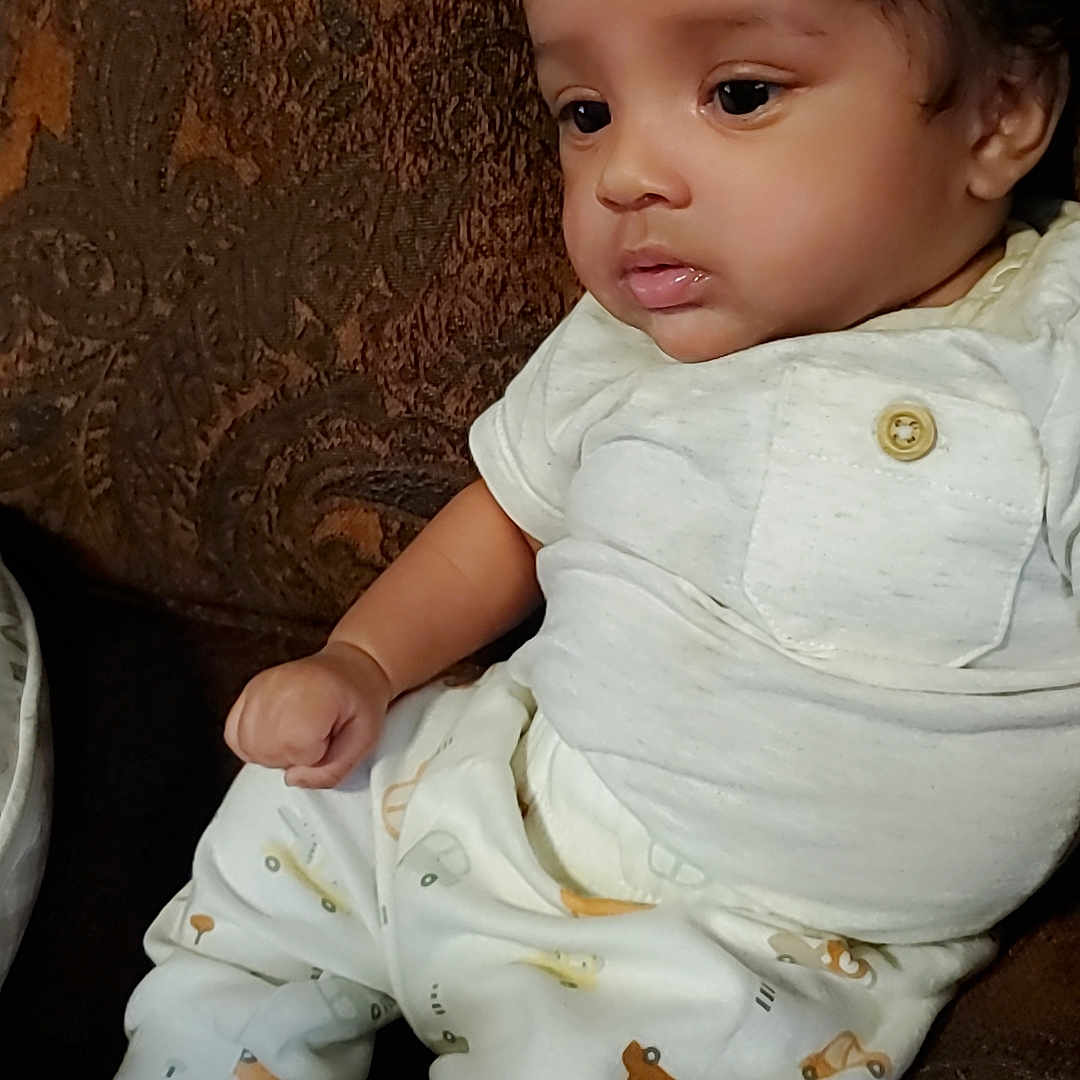 Isley is registered to the contest to win money with this photo: baby, brown_couch, button, child, clothing, couch, curly_hair, cute, face, home, indoor, infant, patterned_pants, portrait, seated, sitting, skin, thoughtful, white_shirt, young_child