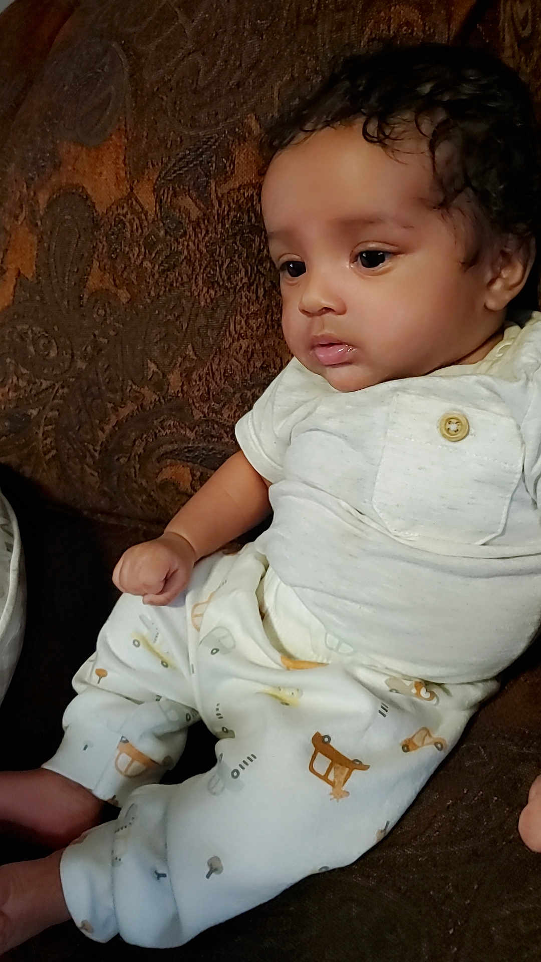 Isley is registered to the contest to win money with this photo: baby, child, curly_hair, white_shirt, patterned_pants, couch, brown_couch, indoor, seated, thoughtful, cute, infant, portrait, clothing, button, face, skin, sitting, young_child, home