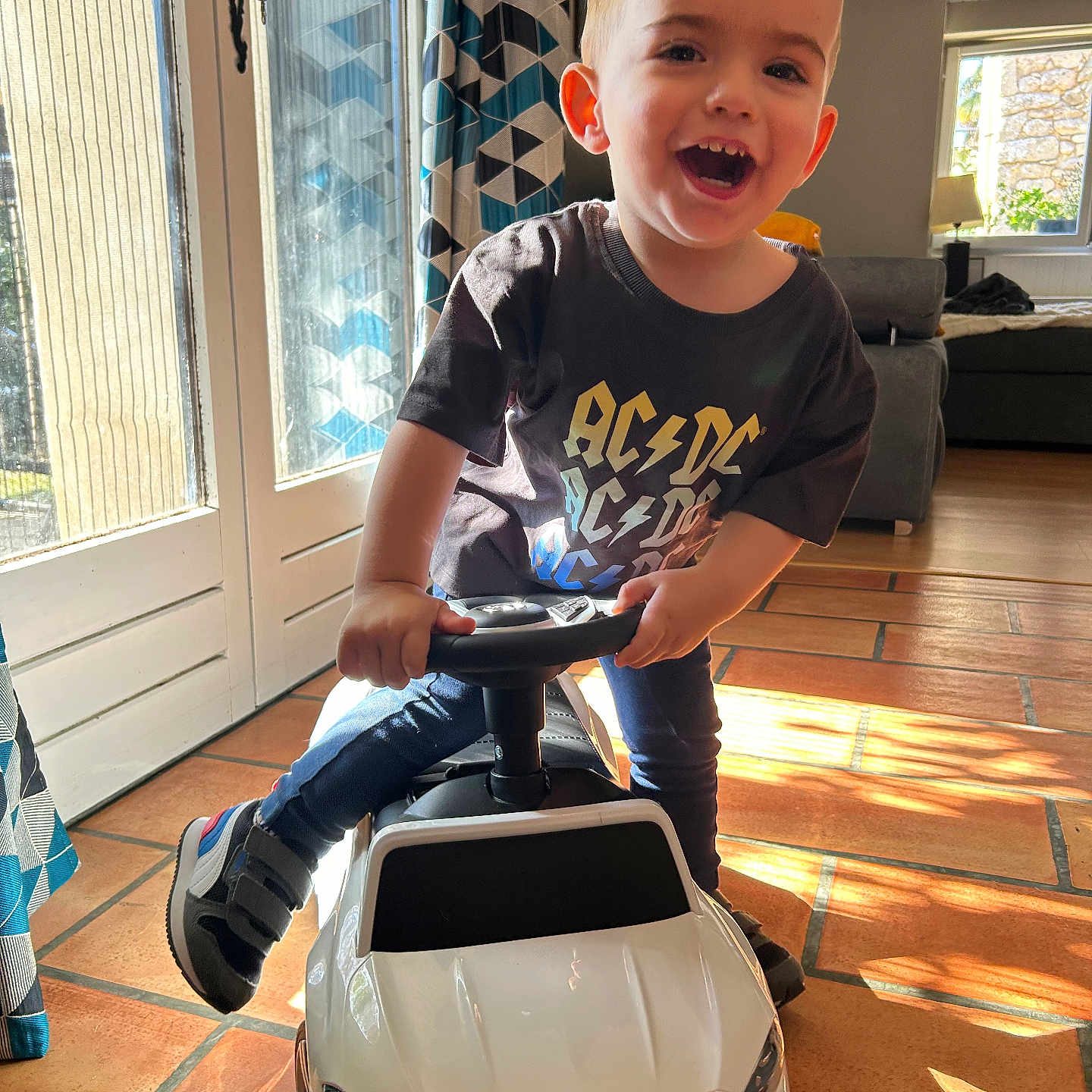Sam participe au concours pour gagner de l'argent avec cette photo : acdc_tshirt, child, curtains, daylight, floor_tiles, footwear, happy, indoor, living_room, miniature_car, person, playful, portrait, short_hair, smiling, sofa, sunlight, toddler, toy_car, window
