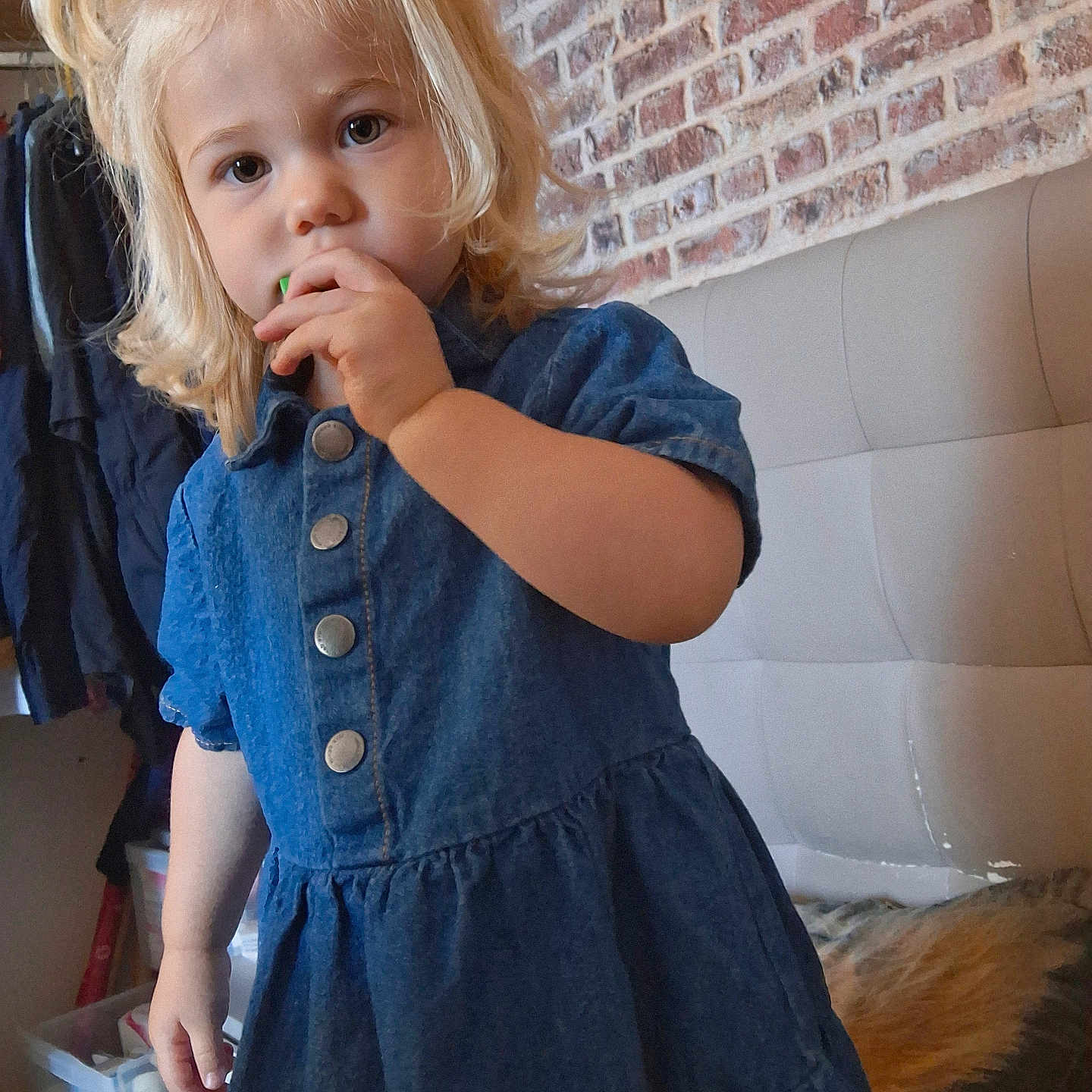 Mya participe au concours pour gagner de l'argent avec cette photo : bed, blonde_hair, brick_wall, child, clothing, coat, curious, curly_hair, denim_dress, face, furniture, hand, headboard, indoor, person, portrait, storage_bins, toddler, toy, young_child