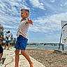 child, boy, barefoot, shorts, shirt, stone_path, outdoor, sky, clouds, water, coast, balance, playful, summer, happy, nature, casual_clothing, person, daytime, landscape