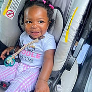 Heaven is registered to the contest to win money with this photo: baby, child, car_seat, smiling, pacifier, hair_ties, pink, white_shirt, plaid_pants, seatbelt, safety_warning, infant, cute, happy, indoors, person, young_child, clothing, car_interior, portrait