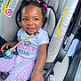baby, child, car_seat, smiling, pacifier, hair_ties, pink, white_shirt, plaid_pants, seatbelt, safety_warning, infant, cute, happy, indoors, person, young_child, clothing, car_interior, portrait