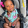 baby, child, smiling, car_seat, pink_bow, floral_dress, bracelet, infant, cute, happy, playing, seatbelt, portrait, indoors, closeup, dark_skin, curly_hair, accessory, baby_toy, seat