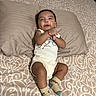 baby, infant, child, smiling, portrait, lying_down, bed, pillow, bedding, patterned_fabric, onesie, socks, bracelet, indoor, home, cute, happy, relaxed, front_view, photography