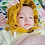 baby, blanket, yellow_bonnet, floral_pattern, soft_texture, infant, portrait, lying_down, cute, child, knitted_clothing, pink_blanket, face, headwear, indoors, calm_expression, person, closeup, colorful_background, warm_clothing