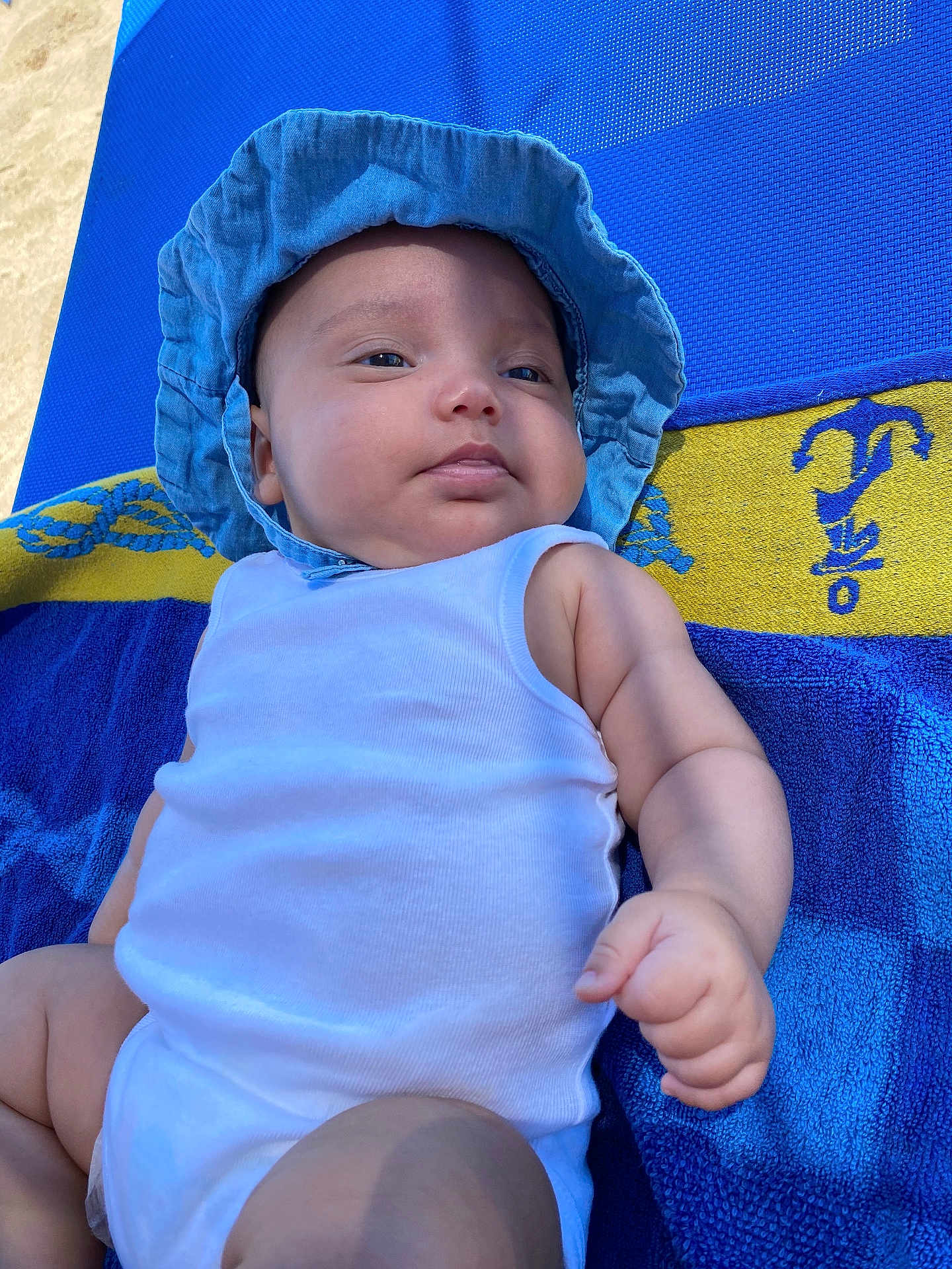 Nyah a rejoint le concours — aidez-le/la à gagner de superbes lots ! baby, infant, blue_hat, white_onesie, towel, blue_towel, yellow_towel, outdoor, sunlight, skin, arm, face, hand, relaxing, summer, child, portrait, cute, chubby, laying_down