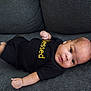 baby, infant, child, couch, sofa, clothing, black_shirt, text, blessed, face, eyes, arms, legs, skin, indoors, furniture, person, lying_down, expression, cute