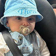 Theo is registered to the contest to win money with this photo: baby, child, car_seat, pacifier, hat, blue_hat, striped_overalls, car_interior, dashboard, seatbelt, infant, clothing, hands, face, eyes, cute, sitting, safety, closeup, indoors