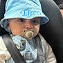 baby, child, car_seat, pacifier, hat, blue_hat, striped_overalls, car_interior, dashboard, seatbelt, infant, clothing, hands, face, eyes, cute, sitting, safety, closeup, indoors