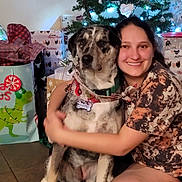 Luna is registered to the contest to win money with this photo: woman, dog, christmas_tree, gift, holiday, smile, indoor, floor, wrapped_presents, decorations, festive, happy, hug, canine, pet, casual_clothing, tile_floor, scarf, friendship, celebration