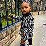 toddler, child, black_and_white_coat, houndstooth, leggings, white_shoes, outdoor, sidewalk, iron_fence, stone_wall, greenery, curious_expression, standing, fashionable, urban, person, portrait, young_child, baby, cute
