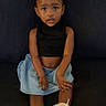 black_top, blue_skirt, brown_skin, casual_clothing, child, couch, curious_expression, cute, dark_background, fashion, footwear, girl, indoor, portrait, short_hair, sitting, small_hands, white_bows, white_sneakers, young_child