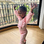 Bella is registered to the contest to win money with this photo: child, toddler, pink_clothing, happy, smiling, indoors, balcony, cityscape, wooden_floor, curtain, railing, window, person, pose, shoes, hair, railing_bars, natural_light, casual, joy