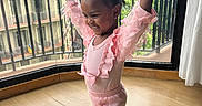 Bella is registered to the contest to win money with this photo: child, toddler, pink_clothing, happy, smiling, indoors, balcony, cityscape, wooden_floor, curtain, railing, window, person, pose, shoes, hair, railing_bars, natural_light, casual, joy
