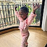 child, toddler, pink_clothing, happy, smiling, indoors, balcony, cityscape, wooden_floor, curtain, railing, window, person, pose, shoes, hair, railing_bars, natural_light, casual, joy
