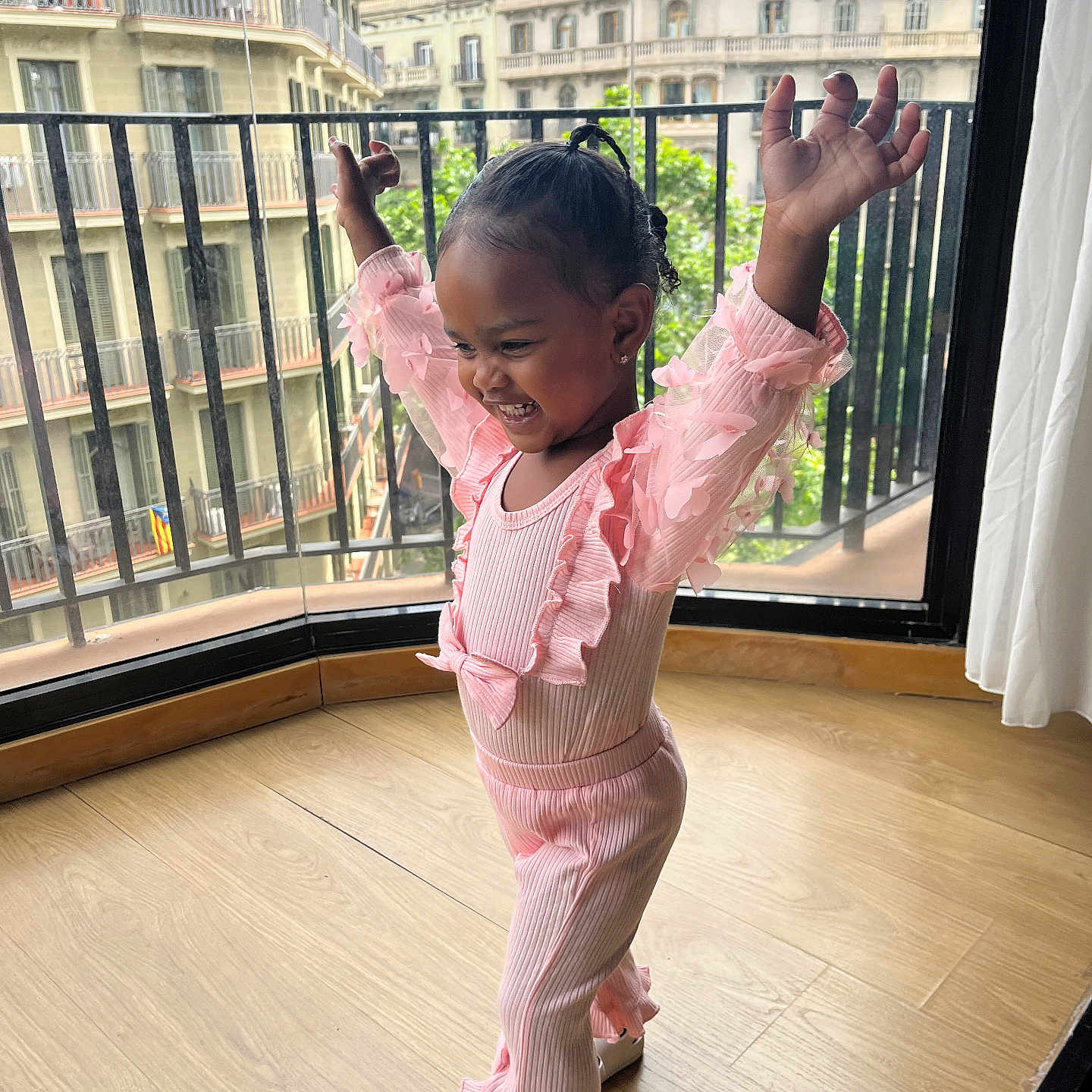 Bella is registered to the contest to win money with this photo: child, toddler, pink_clothing, happy, smiling, indoors, balcony, cityscape, wooden_floor, curtain, railing, window, person, pose, shoes, hair, railing_bars, natural_light, casual, joy