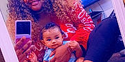 Ka'Hari is registered to the contest to win money with this photo: woman, baby, mirror, selfie, curly_hair, red_shirt, blue_onesie, carpet, couch, television, living_room, smile, holding, sitting, foot, bracelet, phone, reflection, indoor, person