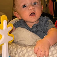 Adonis is registered to the contest to win money with this photo: baby, child, indoor, toy, curious, blue_eyes, cushion, toddler, clothing, hand, face, expression, furniture, background, young_child, playtime, person, infant, head, home
