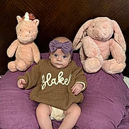 Blakely joined the competition — help win amazing prizes! baby, infant, bow_headband, sweater, plush_unicorn, plush_bunny, stuffed_toy, pillow, purple_bedding, wooden_headboard, bedroom, sitting, legs, feet, wide_eyes, cute_face, knitwear, soft_plush, portrait, cozy