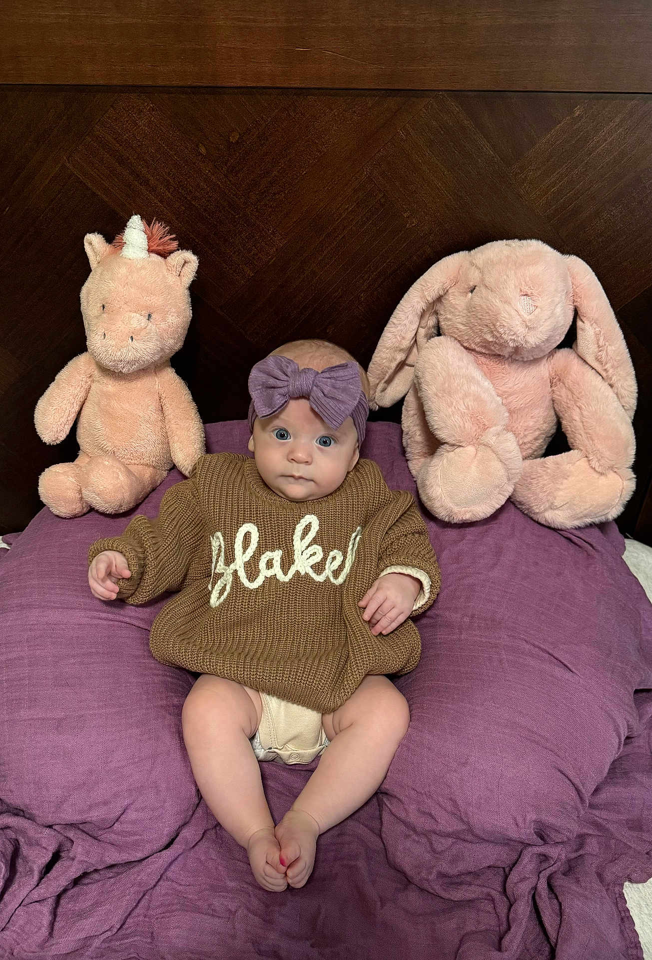 Blakely joined the competition — help win amazing prizes! baby, infant, bow_headband, sweater, plush_unicorn, plush_bunny, stuffed_toy, pillow, purple_bedding, wooden_headboard, bedroom, sitting, legs, feet, wide_eyes, cute_face, knitwear, soft_plush, portrait, cozy