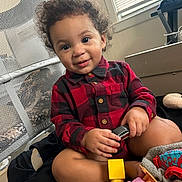 Christian is registered to the contest to win money with this photo: toddler, child, curly_hair, smiling, red_shirt, black_and_red_checkered, toy_car, indoor, bed, window, blinds, mesh_chair, playful, cute, happy, person, skin, sitting, furniture, toy