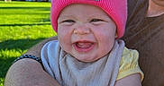 Margaret is registered to the contest to win money with this photo: baby, infant, child, smiling, happy, portrait, pink_hat, beanie, carhartt, outdoor, park, grass, trees, blue_sky, sunlight, adult, parent, cuddling, bib, family