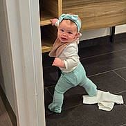 Margaret is registered to the contest to win money with this photo: baby, child, infant, smiling, standing, indoors, cabinet, wooden_furniture, floor, tile_floor, mint_green, headband, bib, long_sleeves, pants, barefoot, doorway, home, portrait, playful