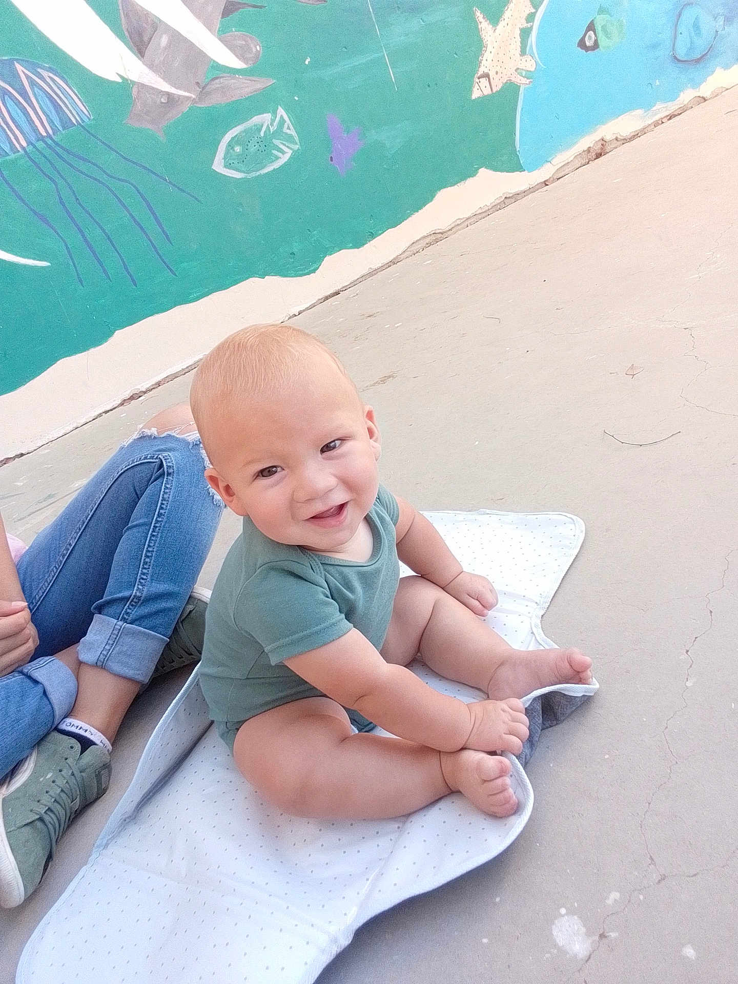 Isaiah joined the competition — help win amazing prizes! baby, child, smile, green_onesie, mat, outdoor, concrete_floor, mural, sea_creatures, person, jeans, sneakers, happy, playful, cute, infant, sitting, casual, colorful, daylight