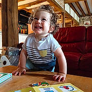 Nino participe au concours pour gagner de l'argent avec cette photo : book, bright, child, clothing, curiosity, curly_hair, furniture, happy, indoor, learning, person, playful, portrait, red_couch, rustic_room, smiling, table, toddler, wood, wooden_beams