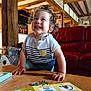 book, bright, child, clothing, curiosity, curly_hair, furniture, happy, indoor, learning, person, playful, portrait, red_couch, rustic_room, smiling, table, toddler, wood, wooden_beams