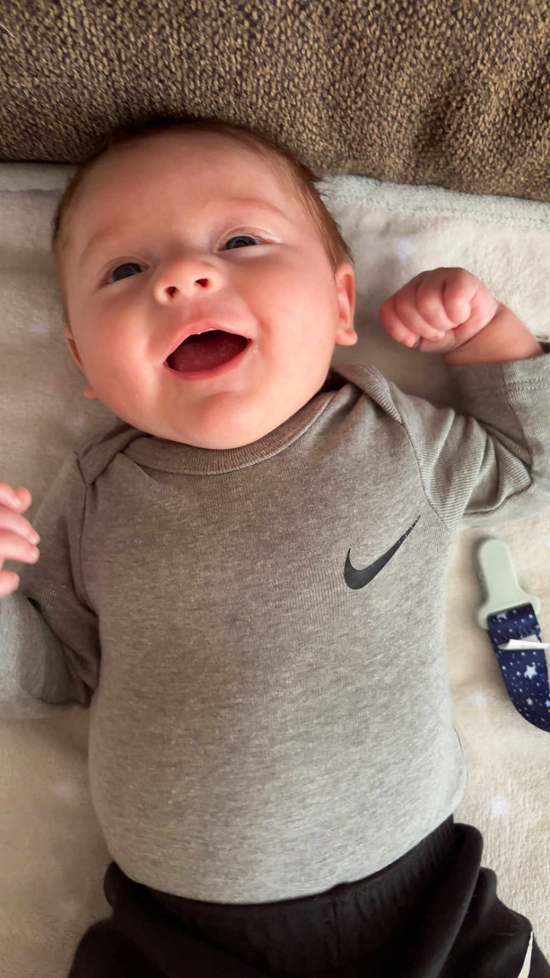 Kylo is registered to the contest to win money with this photo: baby, infant, smile, face, eyes, mouth, onesie, nike_logo, clothing, blanket, pacifier_clip, hand, fist, cheek, lying_down, portrait, closeup, soft_texture, happy, indoor