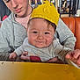 baby, child, teenager, paper_crown, smile, happy, restaurant, booth, orange_seat, table, hands, hug, portrait, family_moment, gray_hoodie, casual_clothing, rosy_cheeks, indoor, person, dining
