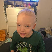 Jackson is registered to the contest to win money with this photo: child, boy, indoor, sitting, green_sweatshirt, hulk, blonde_hair, face, smile, dresser, television, plant, furniture, clothing, room, cozy, casual, person, young, home