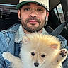 man, puppy, dog, pet, denim_jacket, baseball_cap, new_york_yankees, car_interior, seatbelt, fluffy_dog, cute, portrait, casual_clothing, earbuds, window, daylight, beard, young_adult, holding, closeup