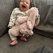 Marnie is registered to the contest to win money with this photo: baby, laughing, stuffed_animal, pink_bunny, couch, gray_couch, onesie, indoor, happy, child, soft_toy, foot, smile, infant, cozy, seated, home, cute, furniture, person