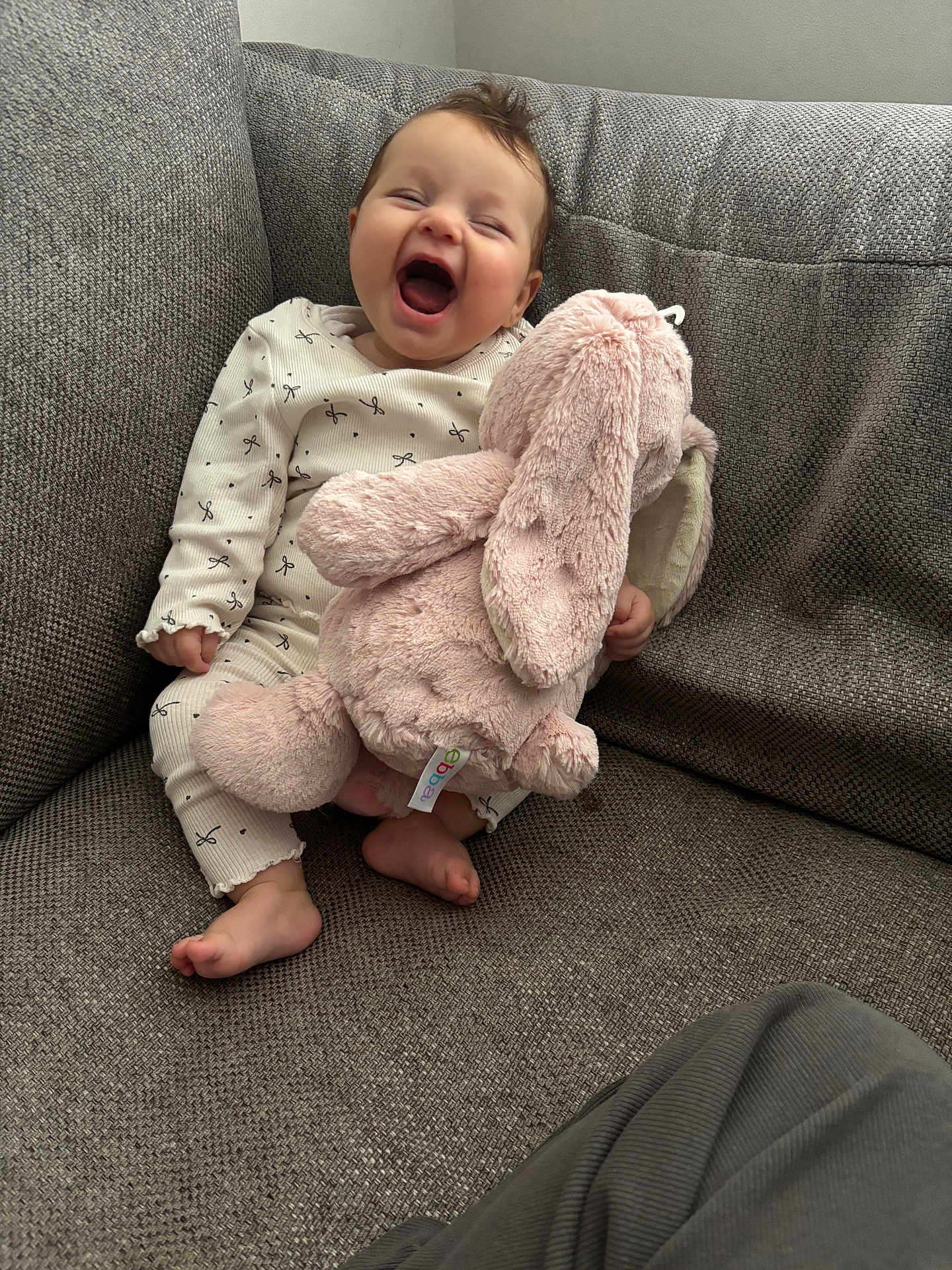 Marnie is registered to the contest to win money with this photo: baby, laughing, stuffed_animal, pink_bunny, couch, gray_couch, onesie, indoor, happy, child, soft_toy, foot, smile, infant, cozy, seated, home, cute, furniture, person