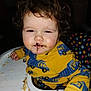 child, toddler, highchair, messy_face, chocolate, food, curly_hair, yellow_clothing, car_pattern, polka_dot, feeding_time, indoor, portrait, baby_food, crumbs, hands, eyes, cheeks, sleepy_expression, candid_photo