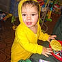 activity_center, baby_snack, blanket, bookcase, brown_eyes, carpet_floor, child, face, hand, hood, indoor, playroom, portrait, red_plate, smile, toddler, toys, waffle, yellow_hoodie, zipper