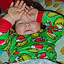 child, toddler, sleeping_child, pajamas, holiday_pajamas, grinch_pattern, hands, closed_eyes, bed, blanket, pillow, face, portrait, cozy, indoors, red_cuffs, messy_hands, smudged_hands, cute, sleepy