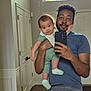 baby, casual_clothing, child, curly_hair, door, floor, greenery, holding, home, indoor, man, mirror, onesie, person, phone, reflection, selfie, smile, socks, window