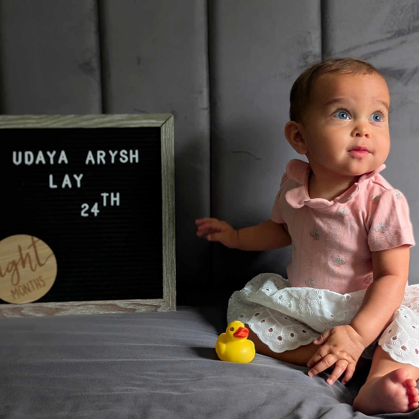 Udaya is registered to the contest to win money with this photo: baby, bed, blue_eyes, celebration, child, cushion, cute, happy, indoors, infant, milestone, person, pink_clothing, portrait, rubber_duck, sign_board, sitting, smiling, soft_lighting, white_skirt