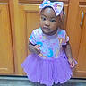 toddler, child, girl, purple_tutu, headband, bow, sandals, kitchen, cabinets, floor, tile_floor, standing, clothing, indoor, person, cute, baby, young_child, footwear, curious