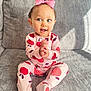 apple_pattern, baby, blue_eyes, child, clapping_hands, cozy, cute, gray_couch, happy, indoor, infant, pajamas, pink_bow, playful, portrait, sitting, smiling, soft_texture, toddler, young_child