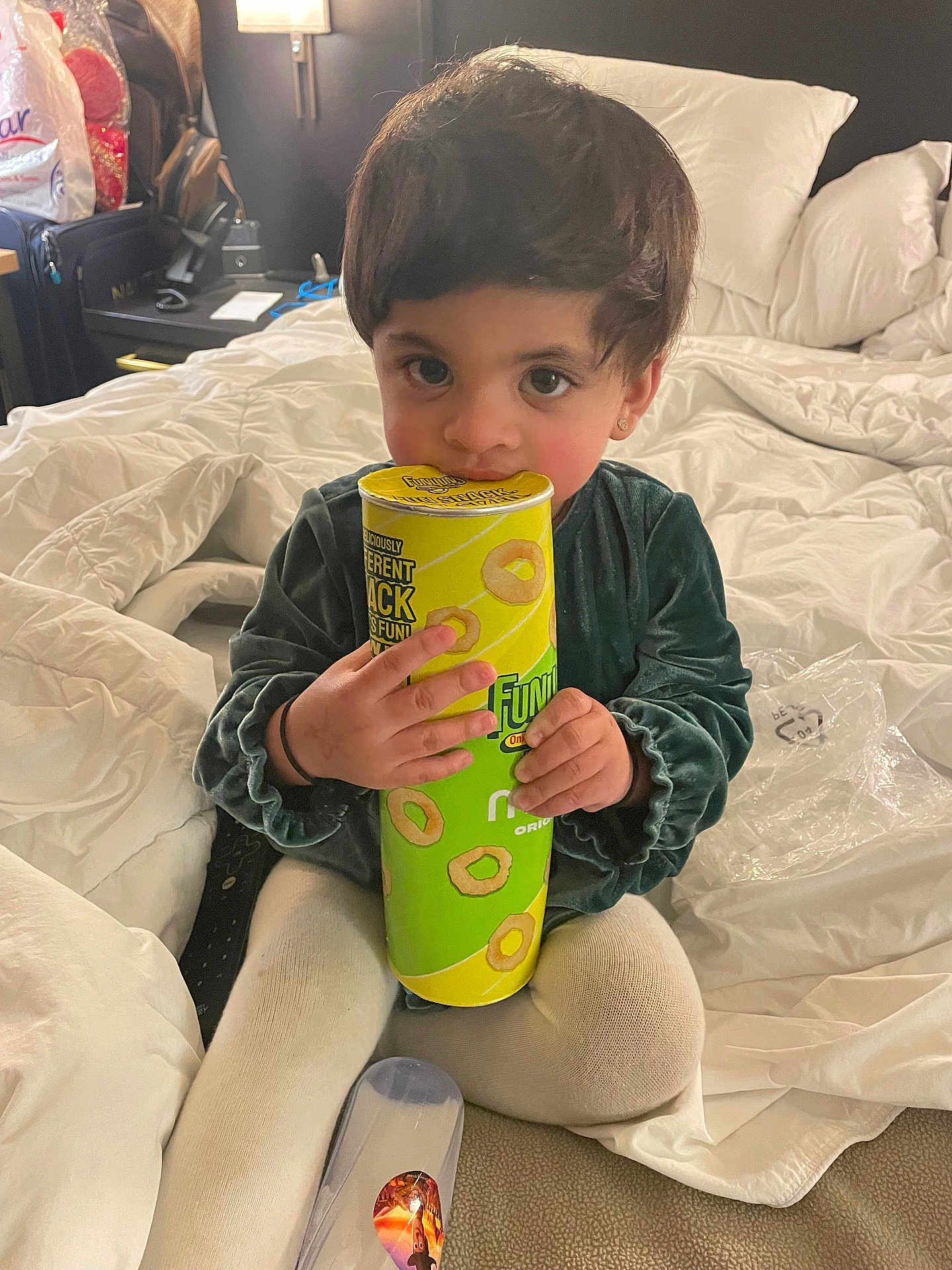 Iman is registered to the contest to win money with this photo: toddler, child, bed, blanket, snack_container, sitting, indoors, curly_hair, cute, clothing, toy, hand, face, eyes, lamp, pillow, carpet, room, plastic_bag, candid