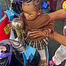 child, toddler, indoor, play_area, carousel, horse, toy, adult_hand, colorful, hair_beads, brown_clothing, plaid_skirt, white_tights, shoes, arcade, floor, fun, curious, ride, entertainment
