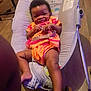 baby, child, smiling, orange_clothing, baby_rocker, indoor, floor, wooden_floor, infant, bracelet, diaper, seat, toy, happy, person, cute, young_child, playful, relaxed, home
