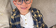 Côme participe au concours pour gagner de l'argent avec cette photo : child, boy, glasses, plaid_shirt, white_shirt, toy_camera, pink, couch, gray_couch, pillow, smile, happy, indoors, casual_clothing, short_hair, person, sitting, hands, digital_screen, home