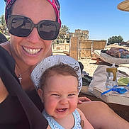 Aaron a rejoint le concours — aidez-le/la à gagner de superbes lots ! woman, baby, smiling, sunglasses, headband, beach, sunhat, tank_top, outdoor, sunny, relaxed, lounge_chair, palm_trees, vacation, water_bottle, goggles, table, sand, man, nap